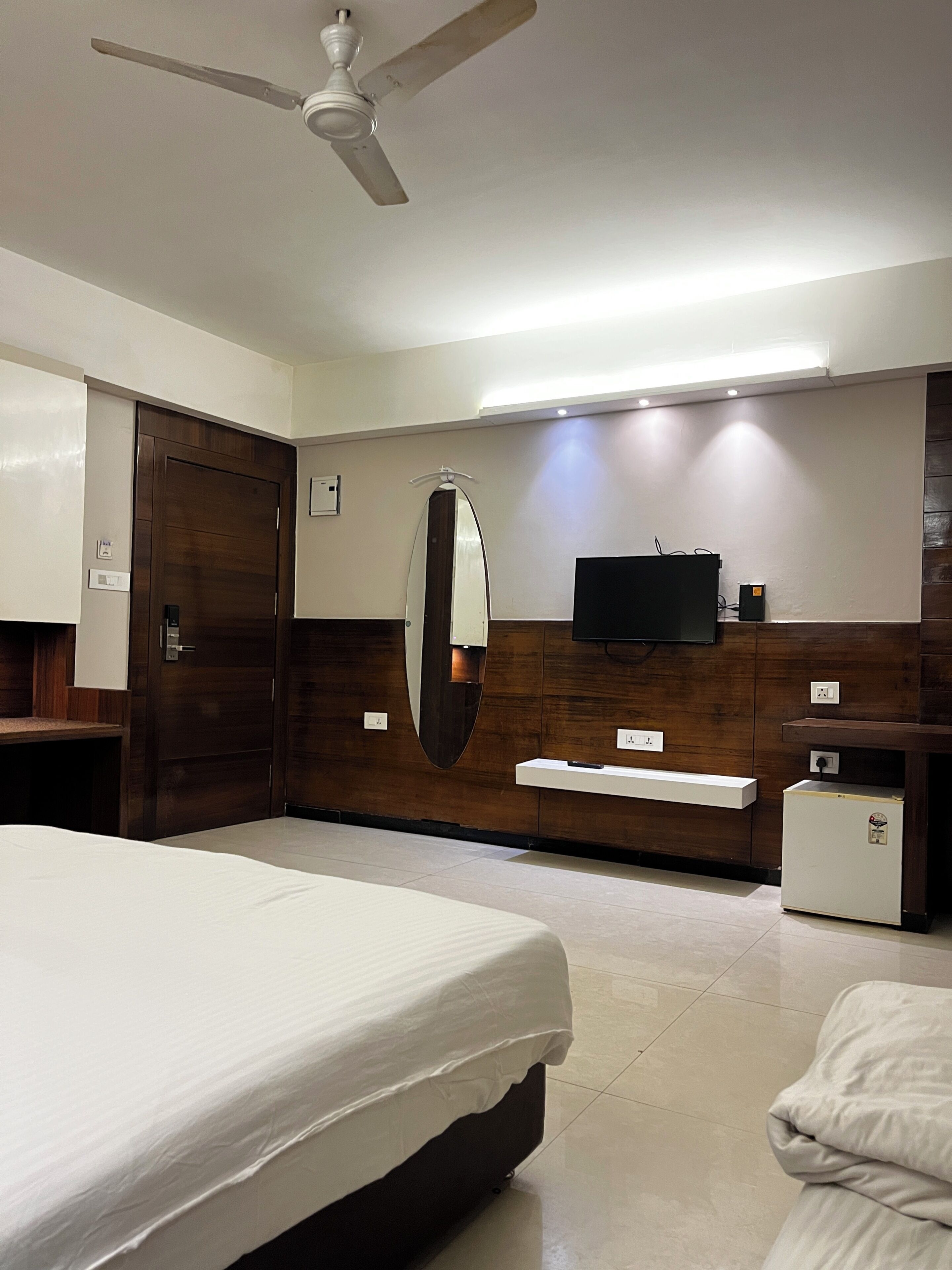Een hotelkamer met een bed, een plafondventilator, een televisie, een spiegel, een kleine koelkast en een houten garderobe.