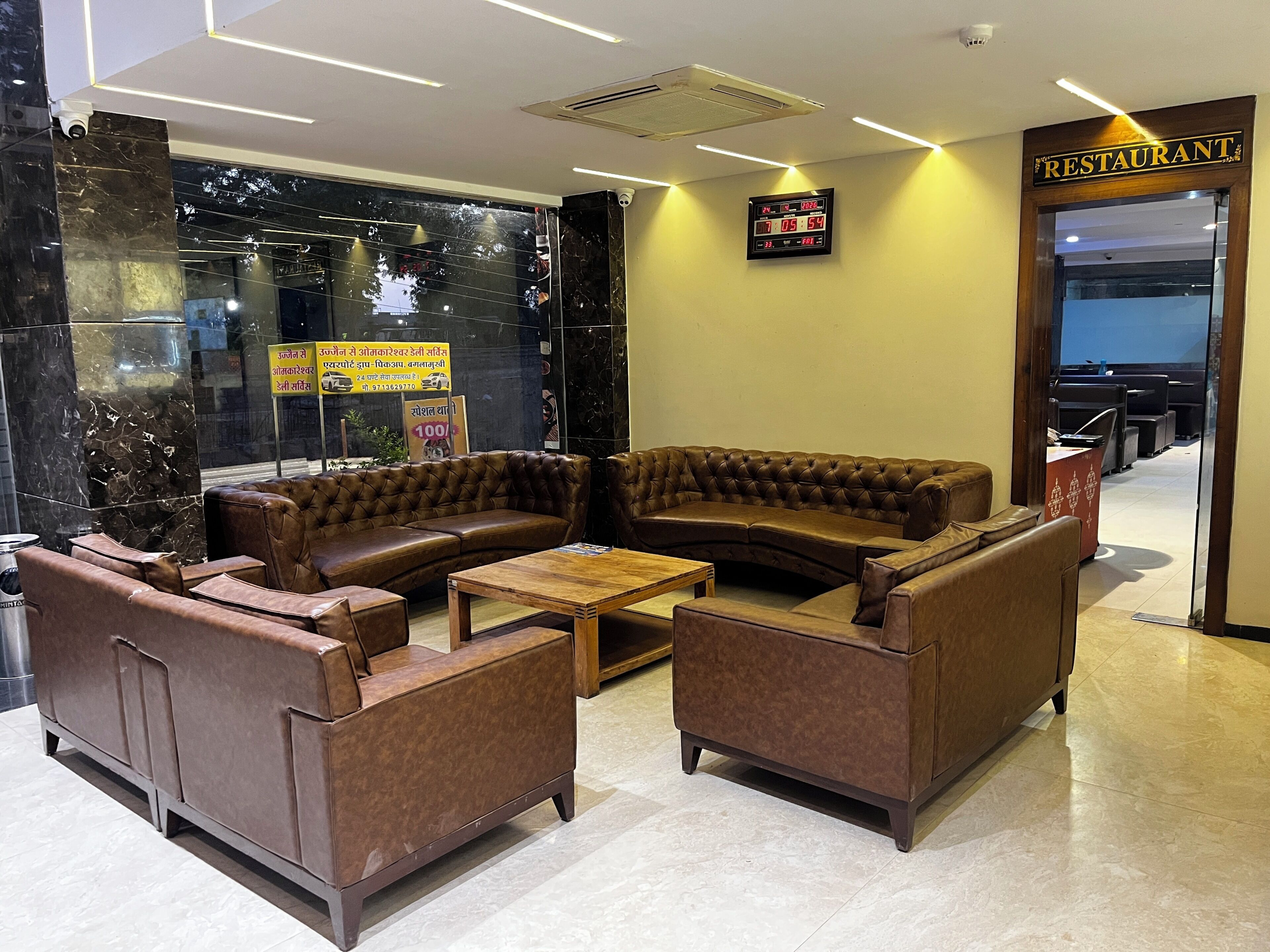 A lobby with a brown leather sofa, two armchairs, a wooden coffee table, a digital clock, and a sign indicating a restaurant.