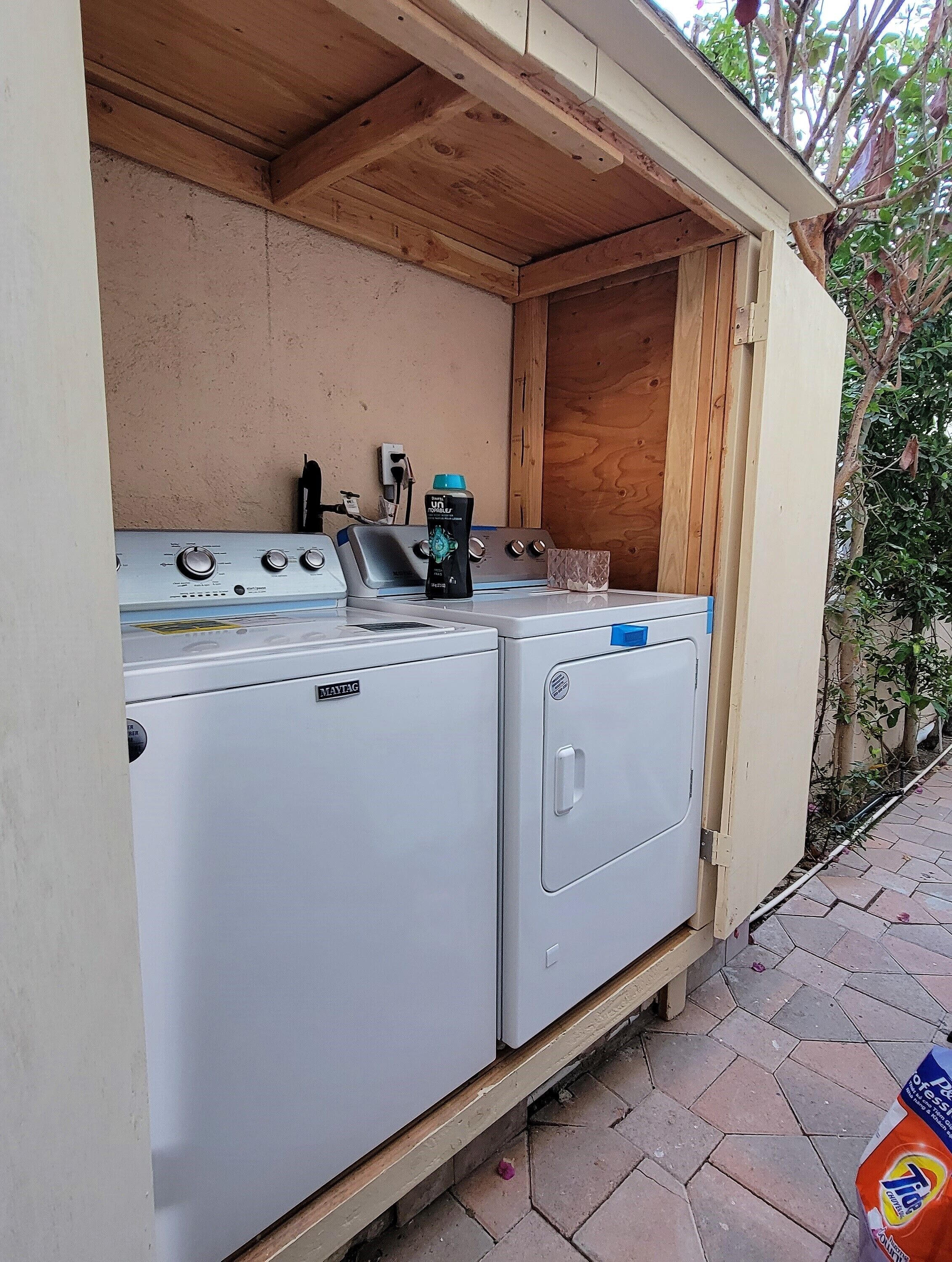 Ein kompakter Waschbereich mit einer Maytag Waschmaschine und Trockner, einem Holzregal und einer Flasche Unilever Geschirrspülmittel.