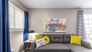 Een moderne woonkamer met een grijze modulaire bank, gele en witte kussens, een klein bijzettafeltje, een lamp en een groot abstract schilderij aan de muur.