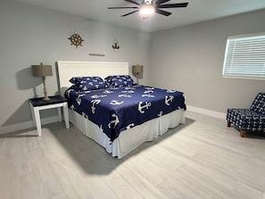 A bedroom with a bed, nightstand, lamp, ceiling fan, wall decor, and a window with blinds.