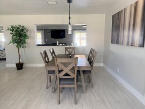 Un coin repas avec une table et des chaises en bois, une plante en pot, un ventilateur de plafond et un téléviseur à écran plat fixé au-dessus d’un îlot de cuisine.