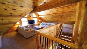 Interior acolhedor de uma cabana de madeira com sofá, televisão e escada de madeira.