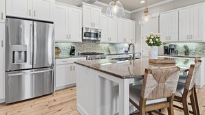 A modern kitchen with white cabinetry, stainless steel appliances, a central island with bar stools, and pendant lighting. - A Wavy Days | 5BR 30A | Steps to Pool | Sleeps 12 | Golf Cart + Bikes (Santa Rosa Beach)