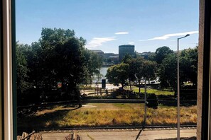 Blick durch ein Fenster auf einen Park mit einem Fluss, Bäumen und einem Gebäude in der Ferne.