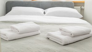 A neatly made bed with pillows and folded towels, a grey headboard, and a window with sheer curtains.