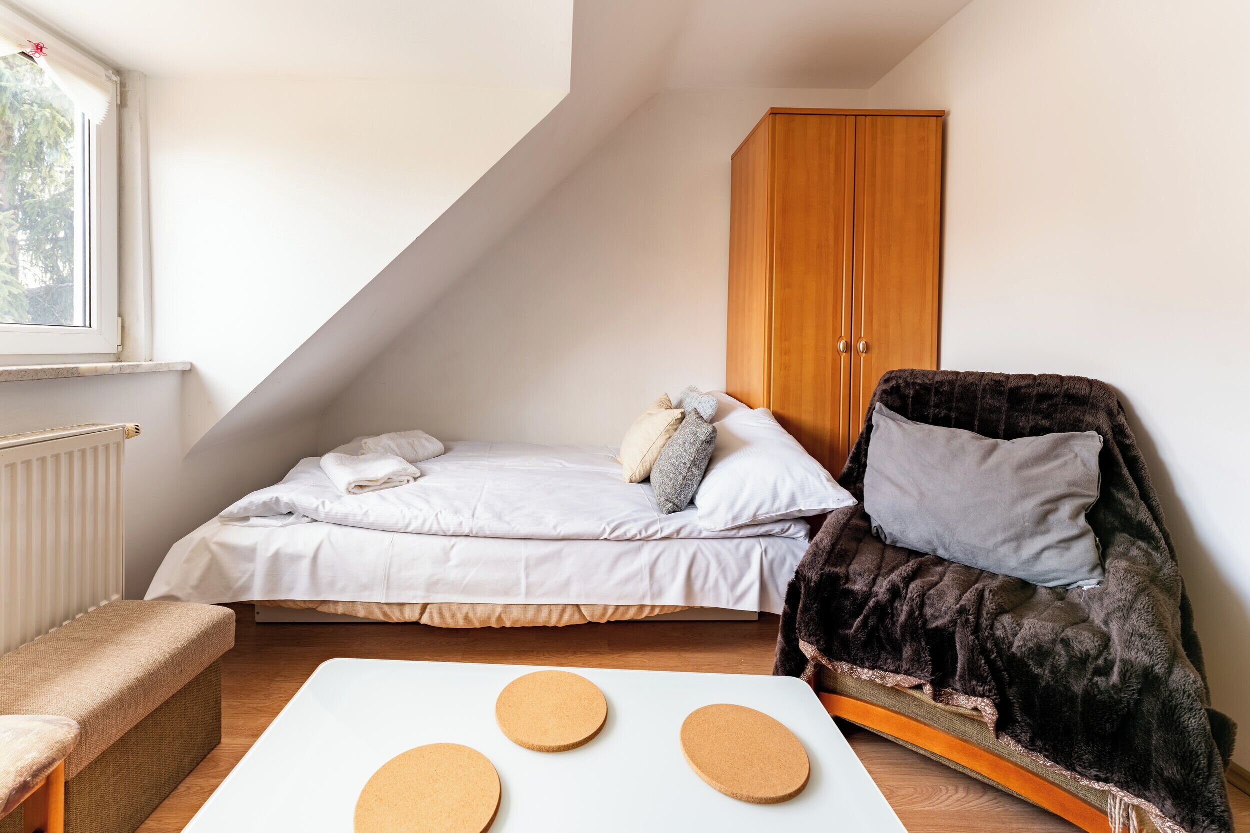 Een kleine, goed verlichte kamer met een bed, een stoel, een kast en een raam.