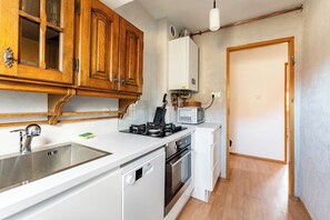 Une cuisine compacte avec des meubles en bois, un four à micro-ondes encastré, une cuisinière à gaz, un lave-vaisselle et un plan de travail blanc.