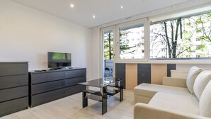 Un soggiorno moderno con un divano, una televisione su un mobile, un tavolino da caffè in vetro e un'ampia finestra con vista su alberi e edifici.