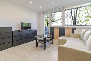 A modern living room with a sofa, a TV on a cabinet, a glass coffee table, and a large window with a view of trees and buildings.
