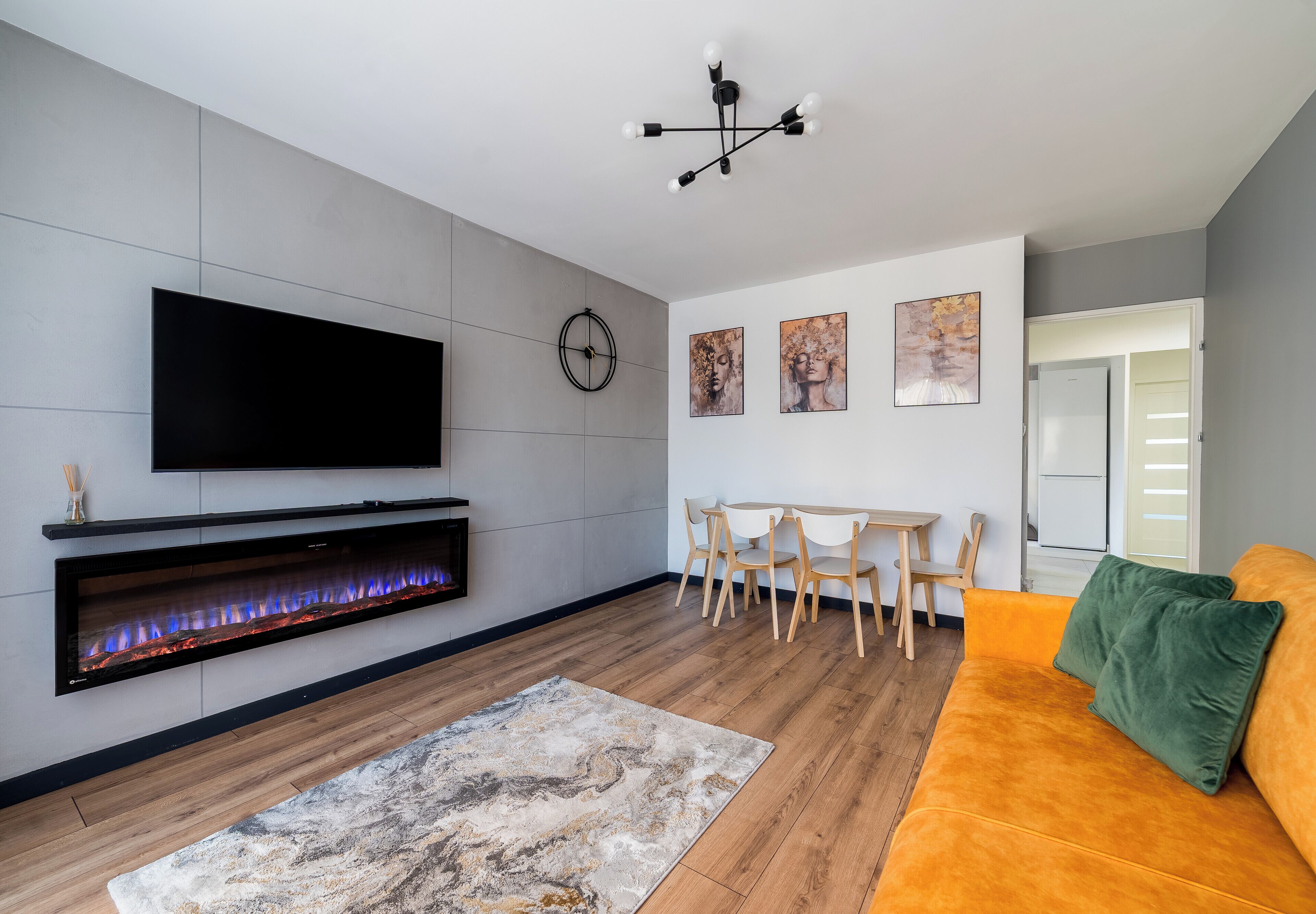 Een moderne woonkamer met een flatscreen televisie, een ingebouwde open haard, een eethoek met een tafel en stoelen, en een bank met kussens.