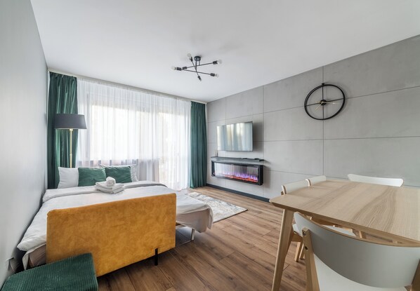 Ein modernes Hotelzimmer mit Bett, Fernseher, Kamin, Esstisch und einem großen Fenster mit Vorhängen.