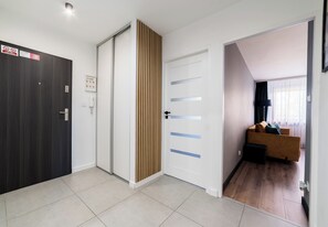 Ein Flur mit einer dunklen Holztür, einer weißen Tür mit blauen Streifen und einem Braunton. Im Raum mit Fenster und Vorhängen steht ein Sofa.