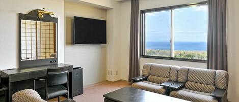 A hotel room with a desk, a sofa, a TV, and a large window with a view of the ocean.