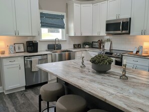 Una cocina moderna con armarios blancos, electrodomésticos de acero inoxidable, encimera de mármol y taburetes de bar.