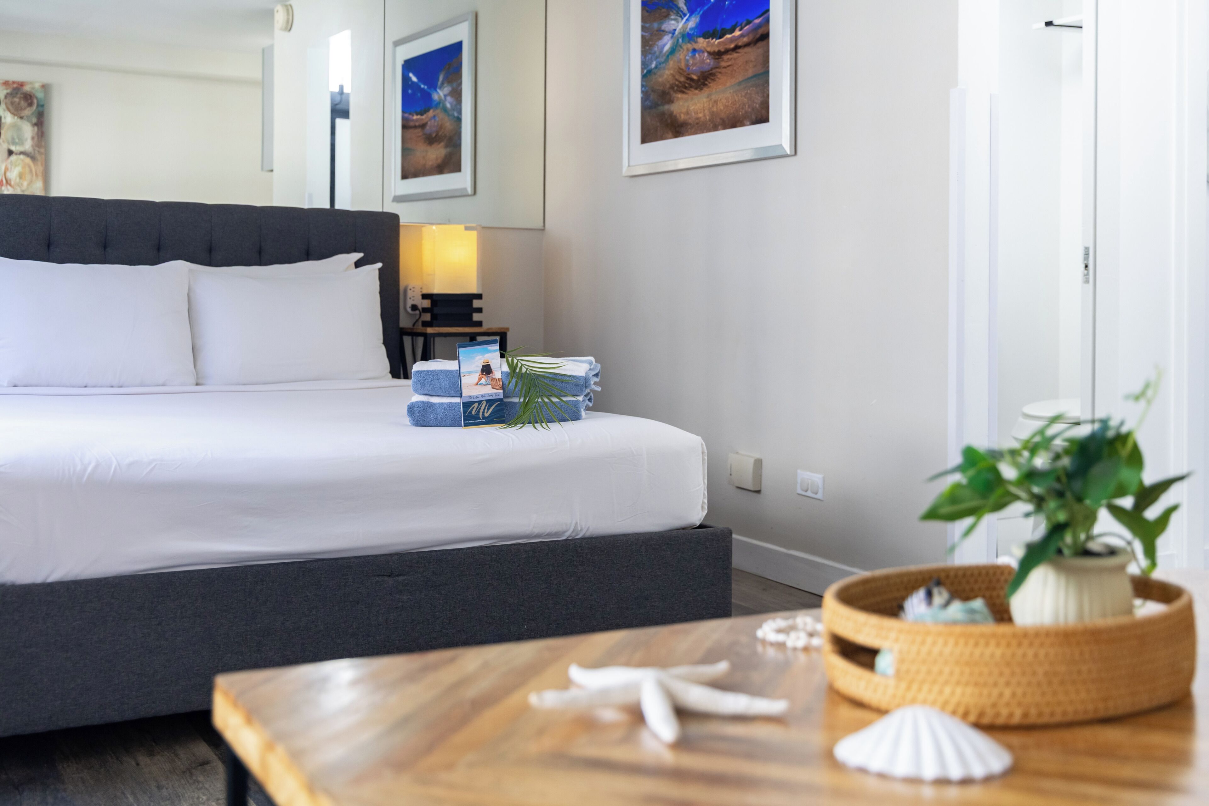 A neatly made bed with white linens, a dark headboard, and a bedside table with a lamp, a framed photo, and a small plant.