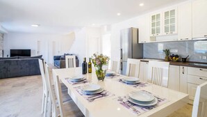 A modern kitchen with white cabinets, a dining area with a long table set for four, and a grey sofa in the background. - Vacation home  by Interhome (Putniković)