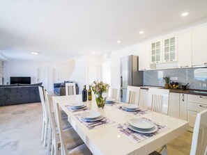 Eine moderne Küche mit weißen Schränken, ein Essbereich mit einem langen Tisch für vier Personen und ein graues Sofa im Hintergrund.