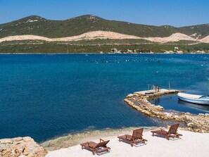 A coastal view with clear blue water, a rocky shoreline, and a row of wooden lounge chairs. - Beachfront Villa Olive Mill with pool by Interhome (Putniković)