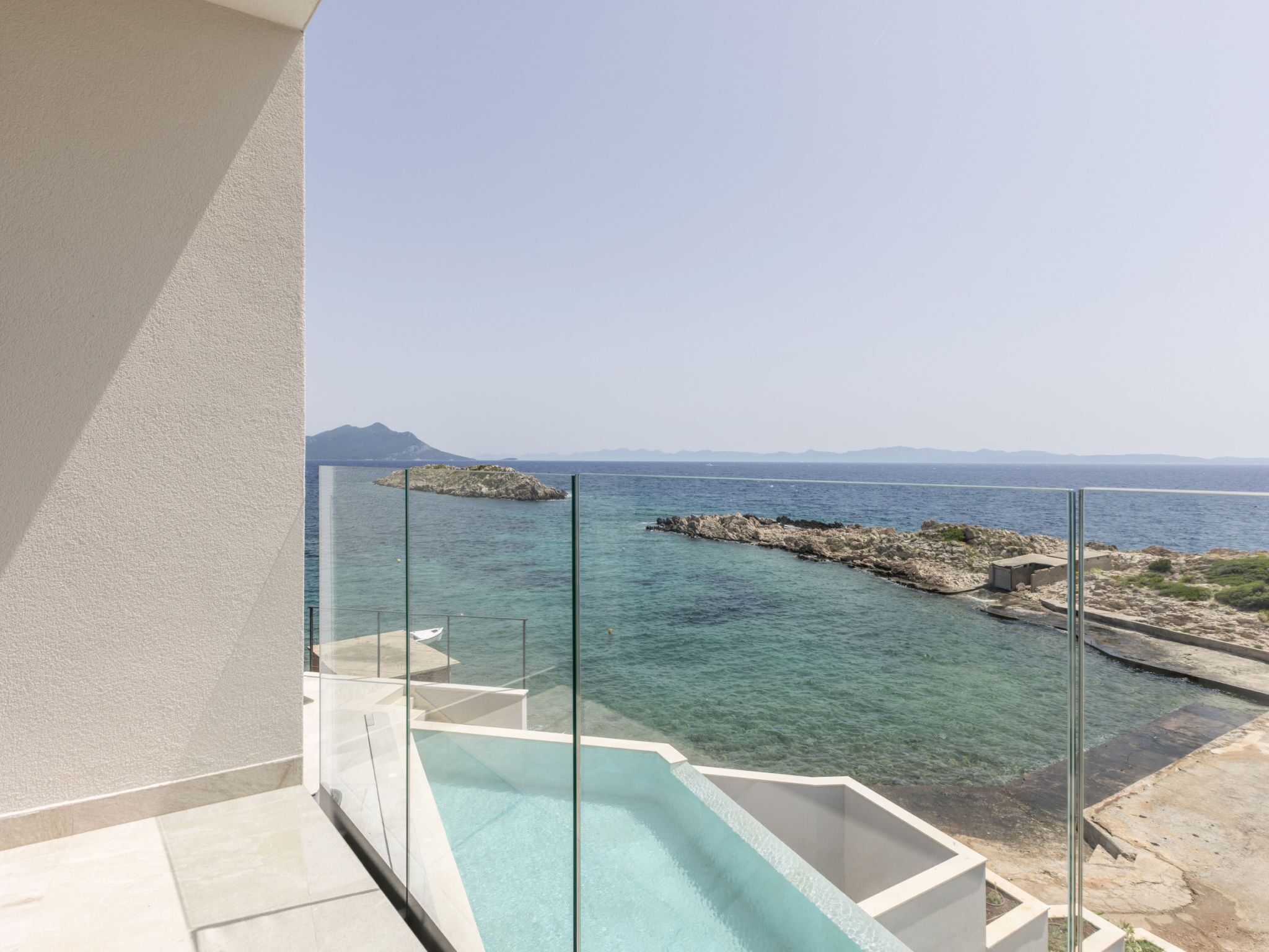 Uma varanda com guarda-corpo de vidro, vista para um mar calmo e formações rochosas.