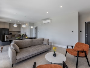 Uma sala de estar moderna com um sofá, uma mesa de centro e uma poltrona laranja. É possível ver uma cozinha ao fundo, com uma mesa de jantar e cadeiras.