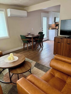 Una sala de estar con un sofá de cuero marrón, una zona de comedor con una mesa y sillas, una cocina con armarios y un televisor, y un equipo de aire acondicionado blanco en la pared.