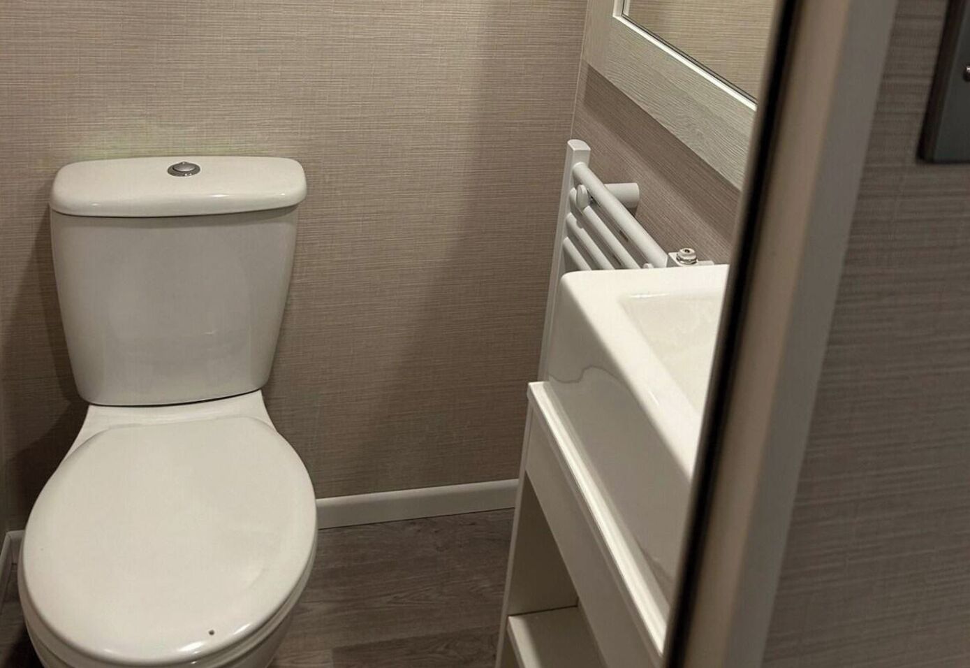 Een wit toilet, een witte radiator en een witte wasbak in een badkamer met beige wanden en een houten vloer.