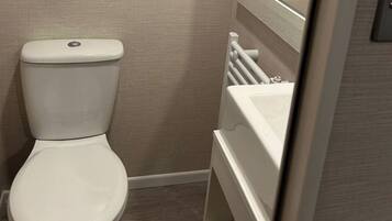 Een wit toilet, een witte radiator en een witte wasbak in een badkamer met beige wanden en een houten vloer.