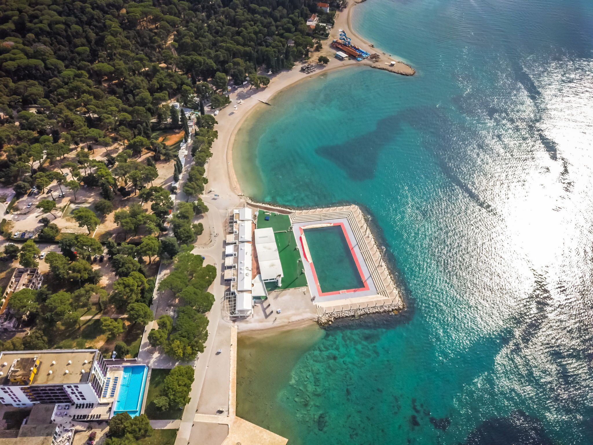 An aerial view of a coastal area with a sandy beach, clear turquoise waters, and a small island with a water park.
