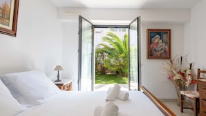 Habitación de hotel con una cama, una mesita de noche, una lámpara, un cuadro y vistas a un jardín.