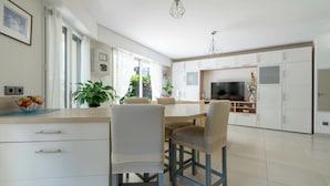 Una cocina moderna con armarios blancos, un comedor con sillas, un televisor y una lámpara colgante.