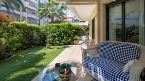 Un balcón con una mesa de vidrio, un sofá a cuadros y vistas a un edificio con balcones y vegetación.
