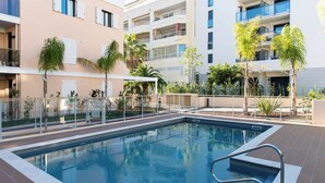 Una piscina con una escalera metálica, rodeada por una cerca y palmeras, con un edificio de varios pisos al fondo.