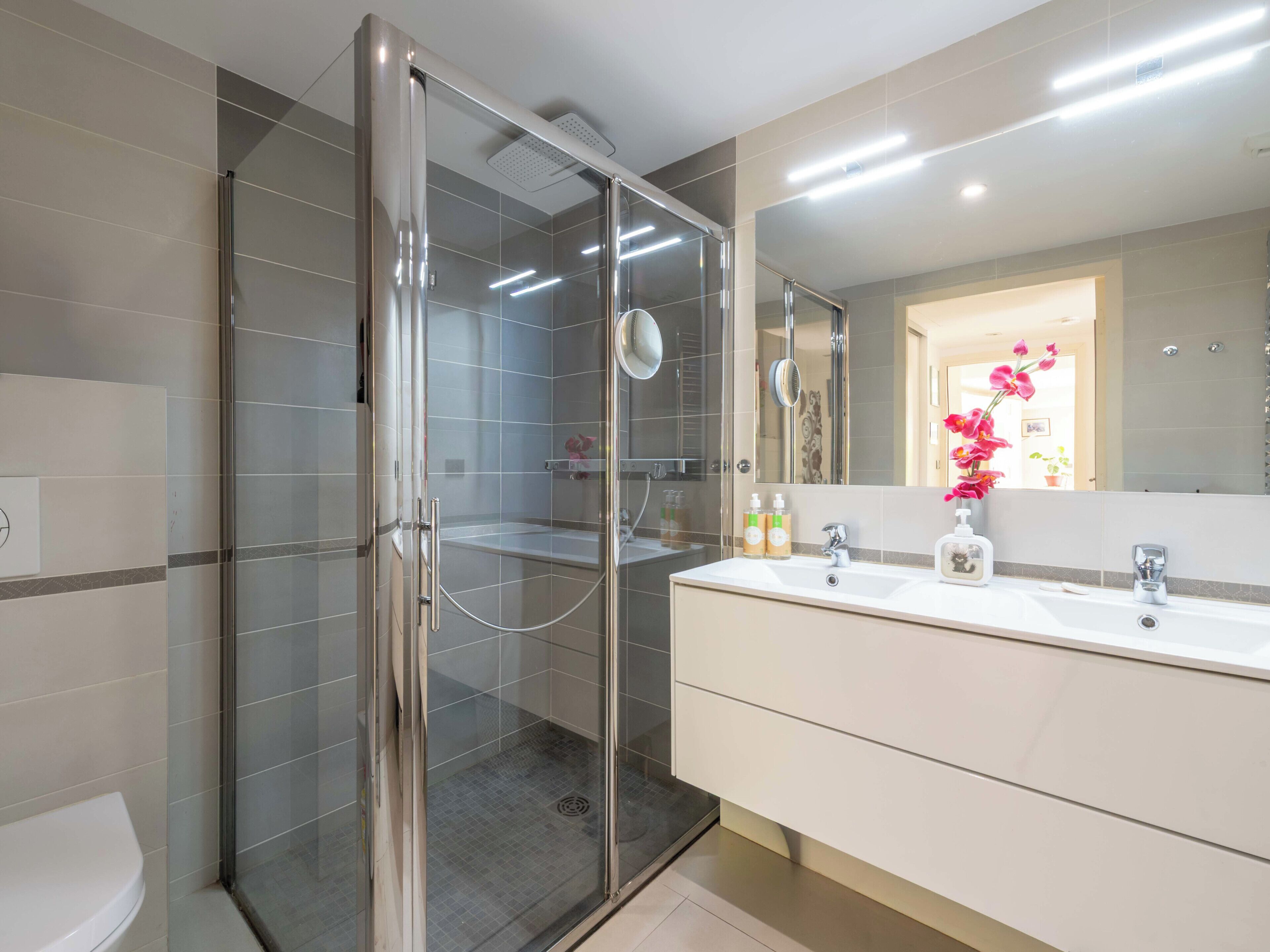 A modern bathroom with a glass-enclosed shower, a white sink with two mirrors, and a beige vanity.