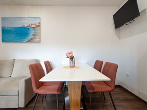A dining area with a white table, six chairs, a vase with a flower, a painting of a coastal scene, and a TV mounted on the wall. - Apartment  by Interhome (Biograd na Moru)