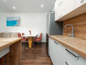 Une cuisine moderne avec des plans de travail en bois, un coin repas avec une table blanche et des chaises rouges, et un tableau représentant un paysage côtier accroché au mur.