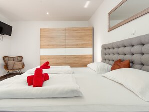 A hotel room with a bed, a wooden wardrobe, a grey upholstered headboard, a wicker chair, and a flat-screen TV mounted on the wall. - Apartment Julia A3, 2+2 by Interhome (Biograd na Moru)
