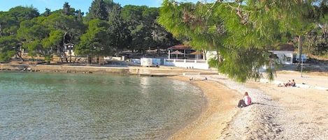Una playa con agua cristalina, piedras y árboles. Hay gente relajándose en la arena.
