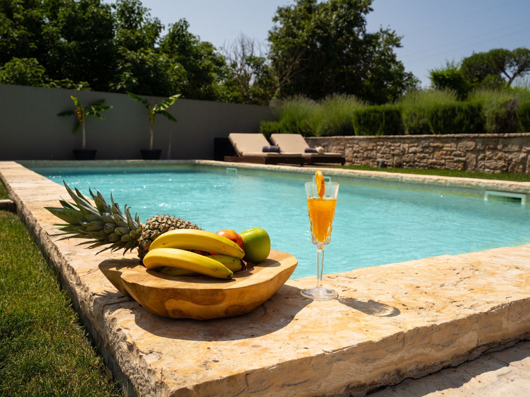 A poolside setting with a fruit bowl and a glass of orange juice on a stone ledge.