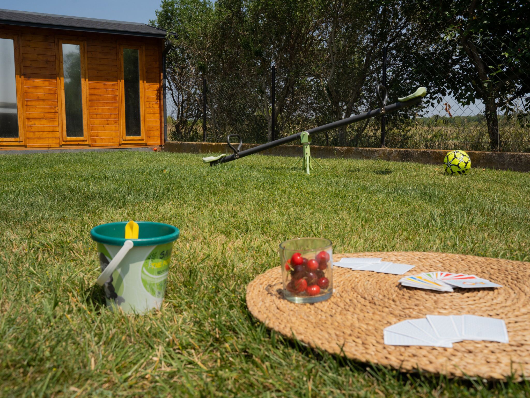 A grassy outdoor area with a wooden building, a metal swing set, a yellow ball, a bucket with a straw, and a glass with cherries.