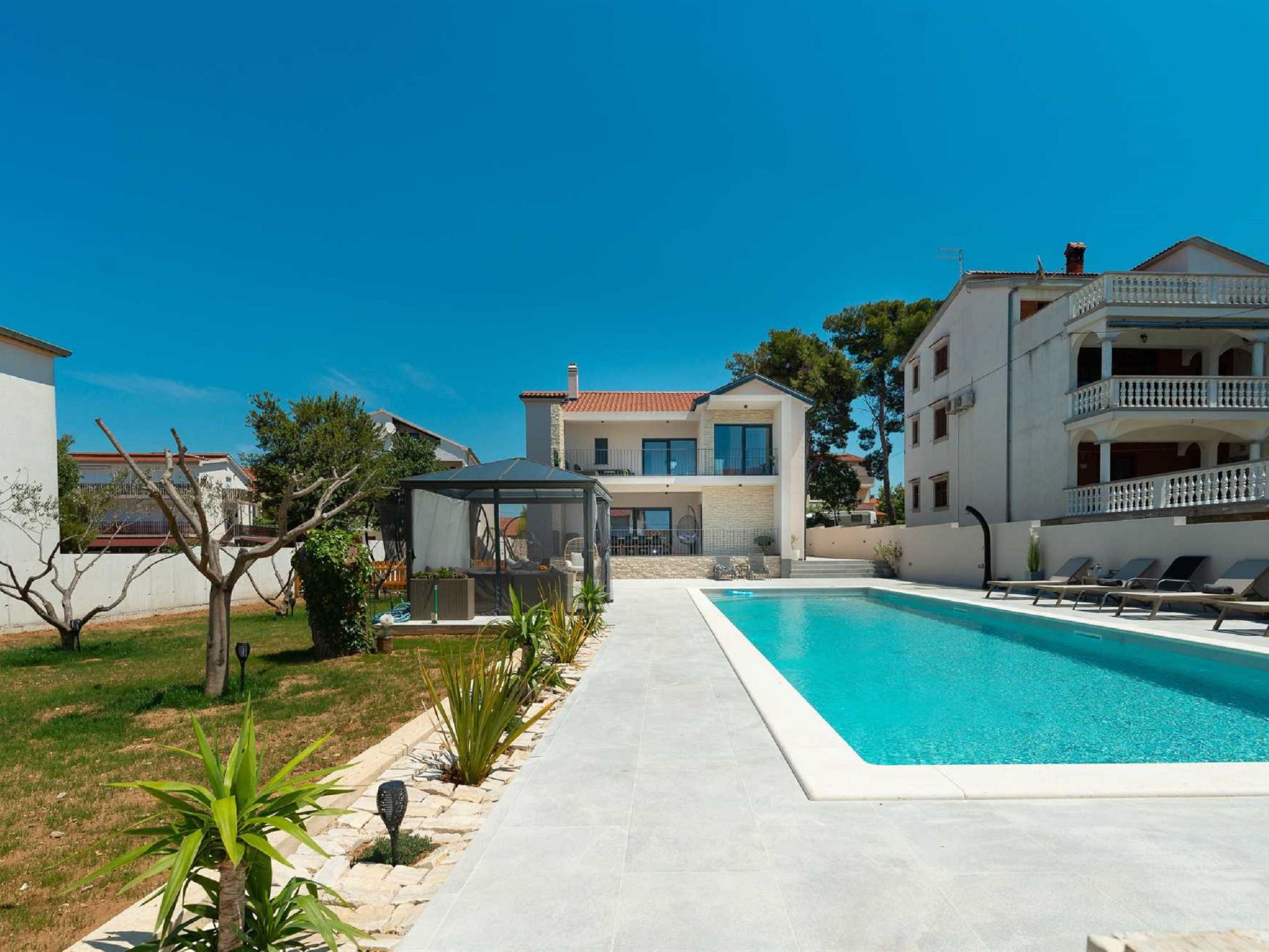 A swimming pool with a clear blue water, surrounded by a paved walkway, and adjacent to a building with a balcony.