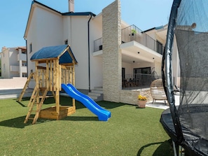 Ein modernes Haus mit einem hölzernen Spielhäuschen und einer blauen Rutsche auf Kunstrasen.