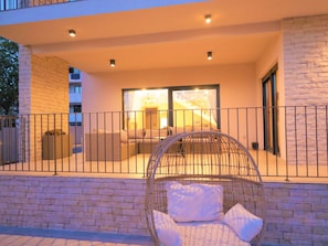 Un balcon avec un fauteuil en osier et des coussins, un mur en pierre et une vue sur l’intérieur, visible à travers une porte vitrée.