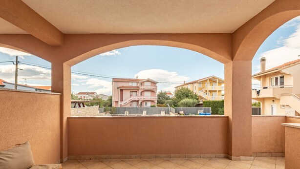 Ein überdachter Außenbereich mit Holztisch und einer Couch, Blick auf Gebäude im Hintergrund und einem klaren Himmel.