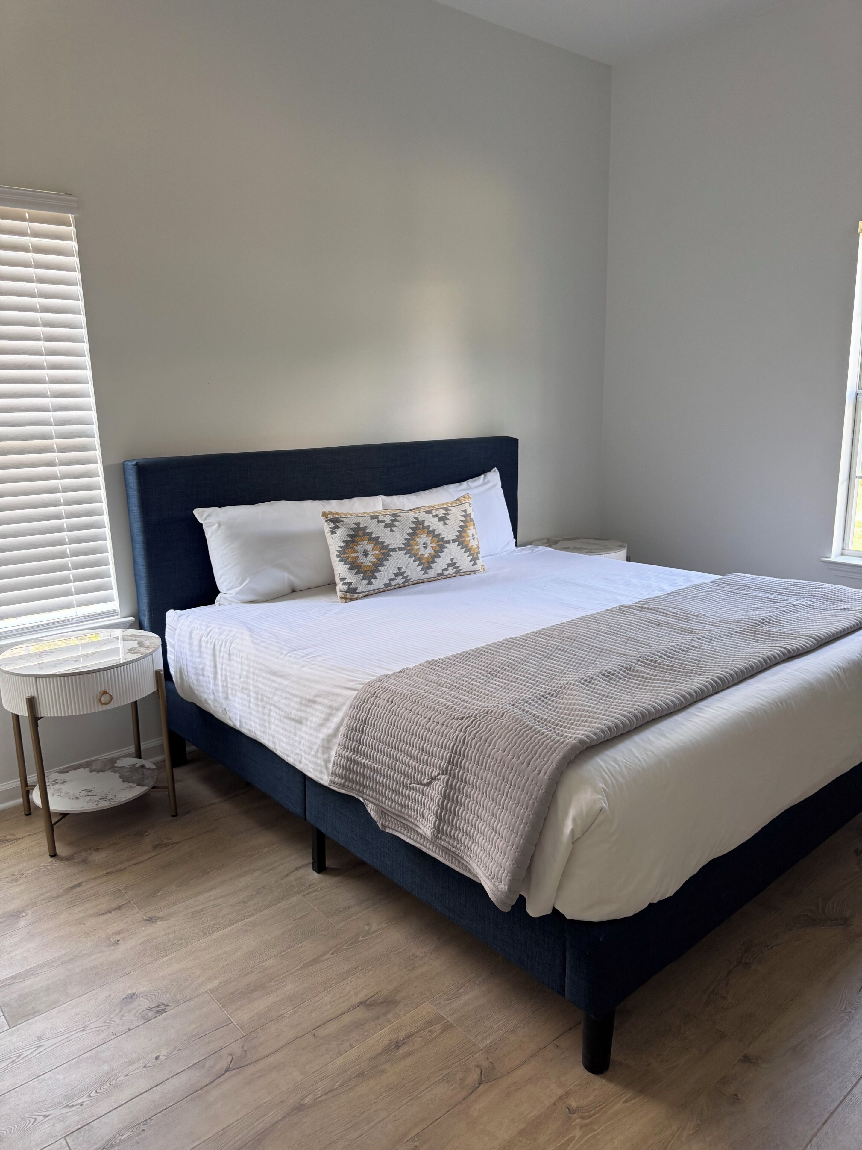 A neatly made bed with a dark headboard, a round bedside table, and a window with blinds.