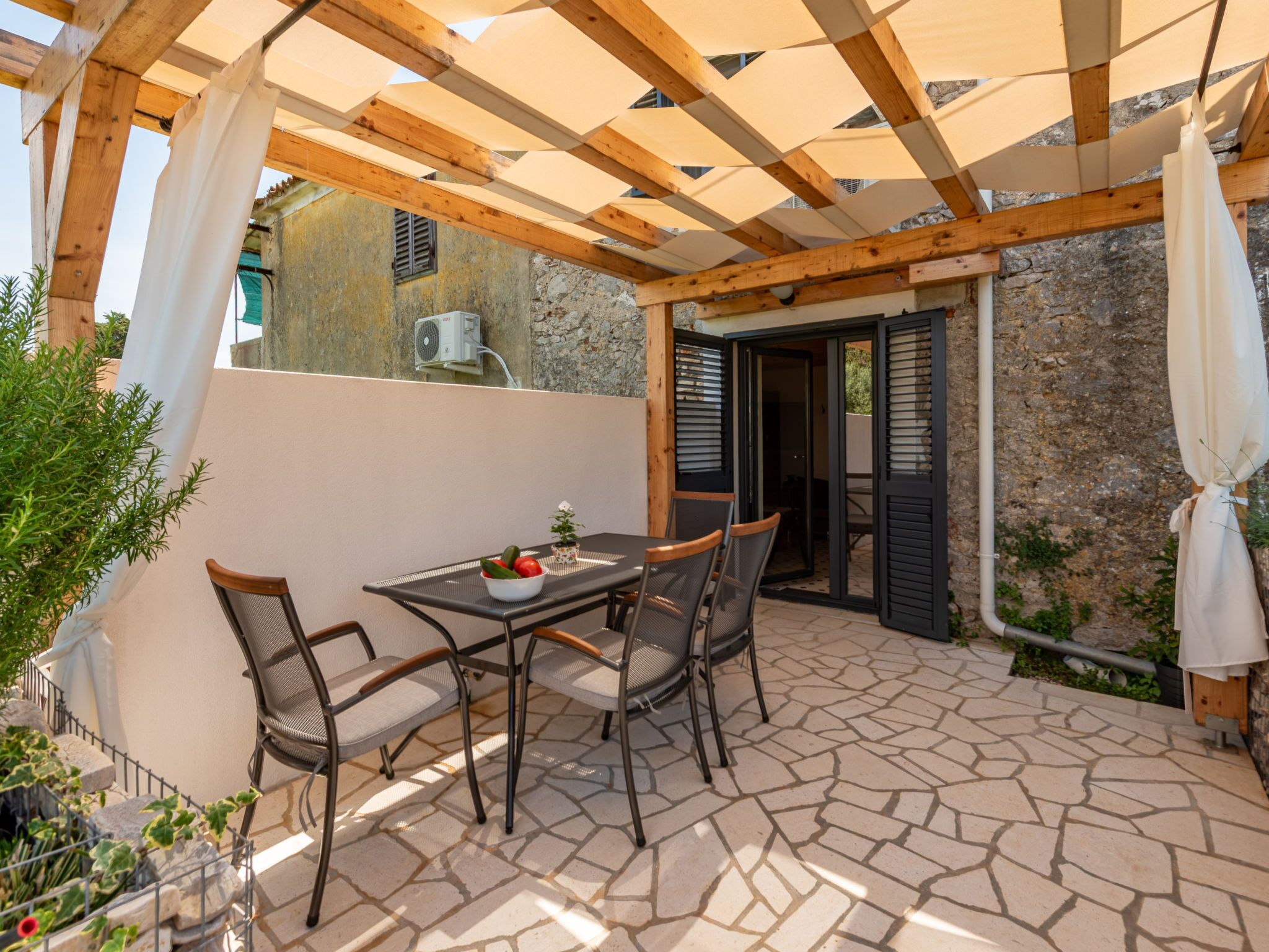 Um pátio externo com mesa, cadeiras, estrutura de pérgola e piso de pedra.