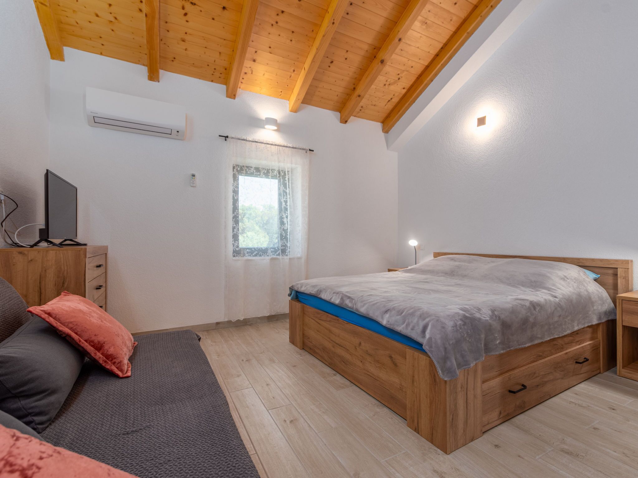 A bedroom with a wooden bed, a TV on a stand, a window with a curtain, and a ceiling with exposed beams.