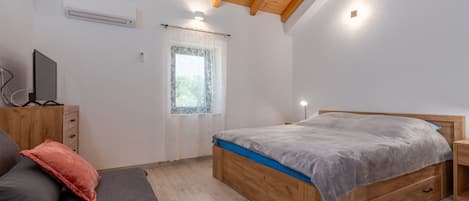 A bedroom with a wooden bed, a TV on a stand, a window with a curtain, and a ceiling with exposed beams.