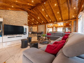 Ein geräumiger Wohnbereich mit Steinwand, Holzbalkendecke, einem Flachbildfernseher, einem grauen Sofa mit roten Kissen und einem Essbereich im Hintergrund.