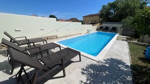 Ein rechteckiger Pool mit gefliestem Sonnendeck, umgeben von einer niedrigen weißen Mauer und einer Steinfassung. Zwei Liegestühle und ein kleiner Tisch mit Glasplatte sind vorhanden.